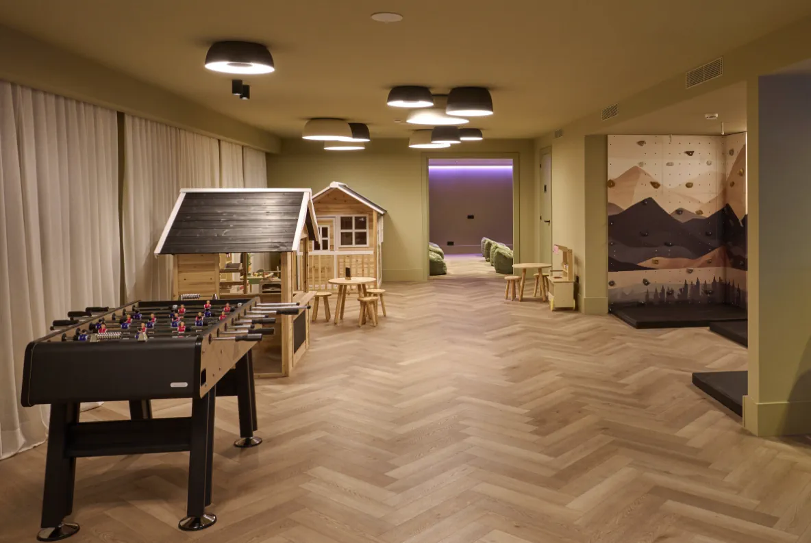 Kids club de Casa Serras amb casetes de fusta, futbolí i rocòdrom infantil sobre parquet en espiga.